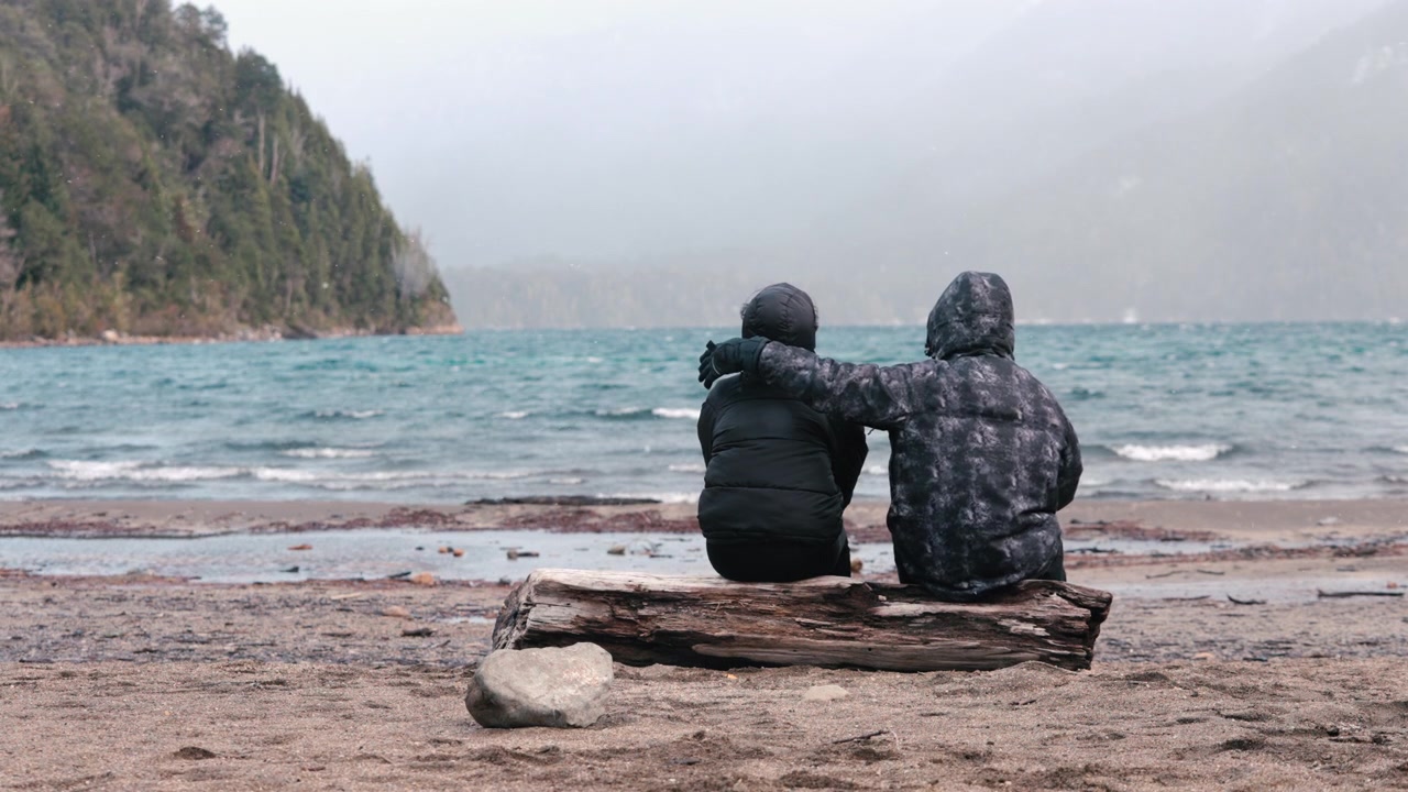 Two individuals sit on a log by the sea, one comforting the other with an arm around their shoulders. The scene captures a sense of support and companionship. Suitable for themes of friendship or comf