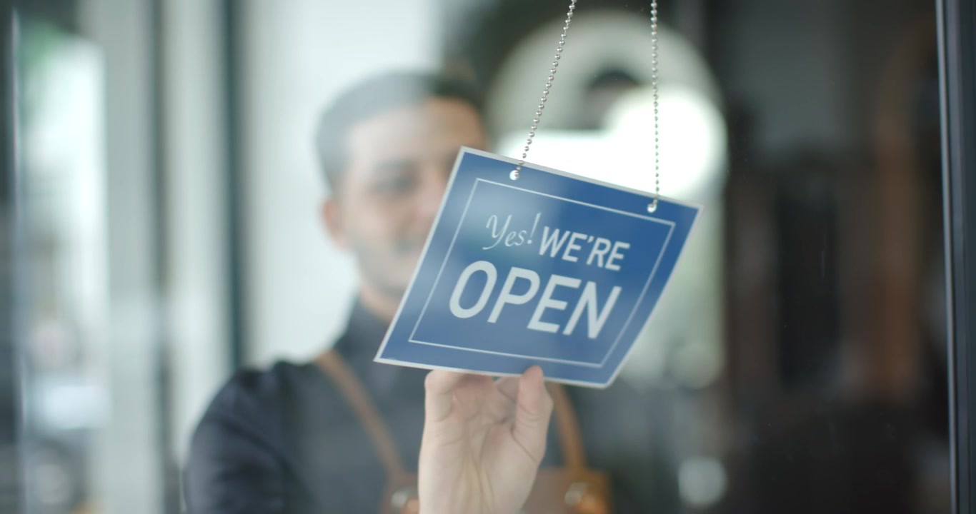 African American apron-clad man reaching, flipping red CLOSED sign on chain at glass door to open
