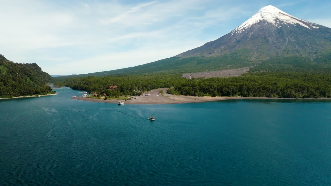 Drone establishing dolly across turquoise lake with surrounding mountains near Petrohue Port, Chile with snowcapped mountain and boats in water