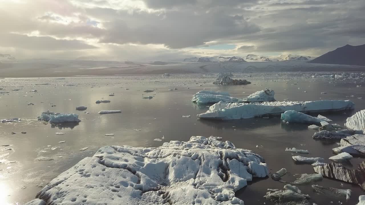Aerial truck right of ice bocks floating in JÃ¶kulsÃ¡rlÃ³n glacial lake, BreiÃ°amerkurjÃ¶kull glacier in background on a cloudy day, VatnajÃ¶kull National Park, Iceland