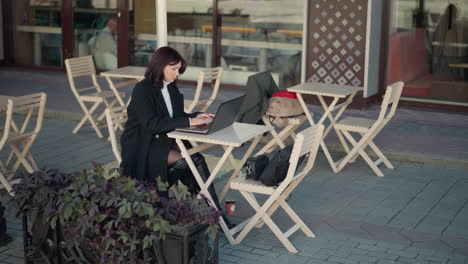 mujer joven de cabello oscuro trabajando en una computadora portátil al aire libre en un ambiente acogedor de centro comercial, está escribiendo en una laptop en una mesa rodeada de sillas y plantas, con el fondo borroso