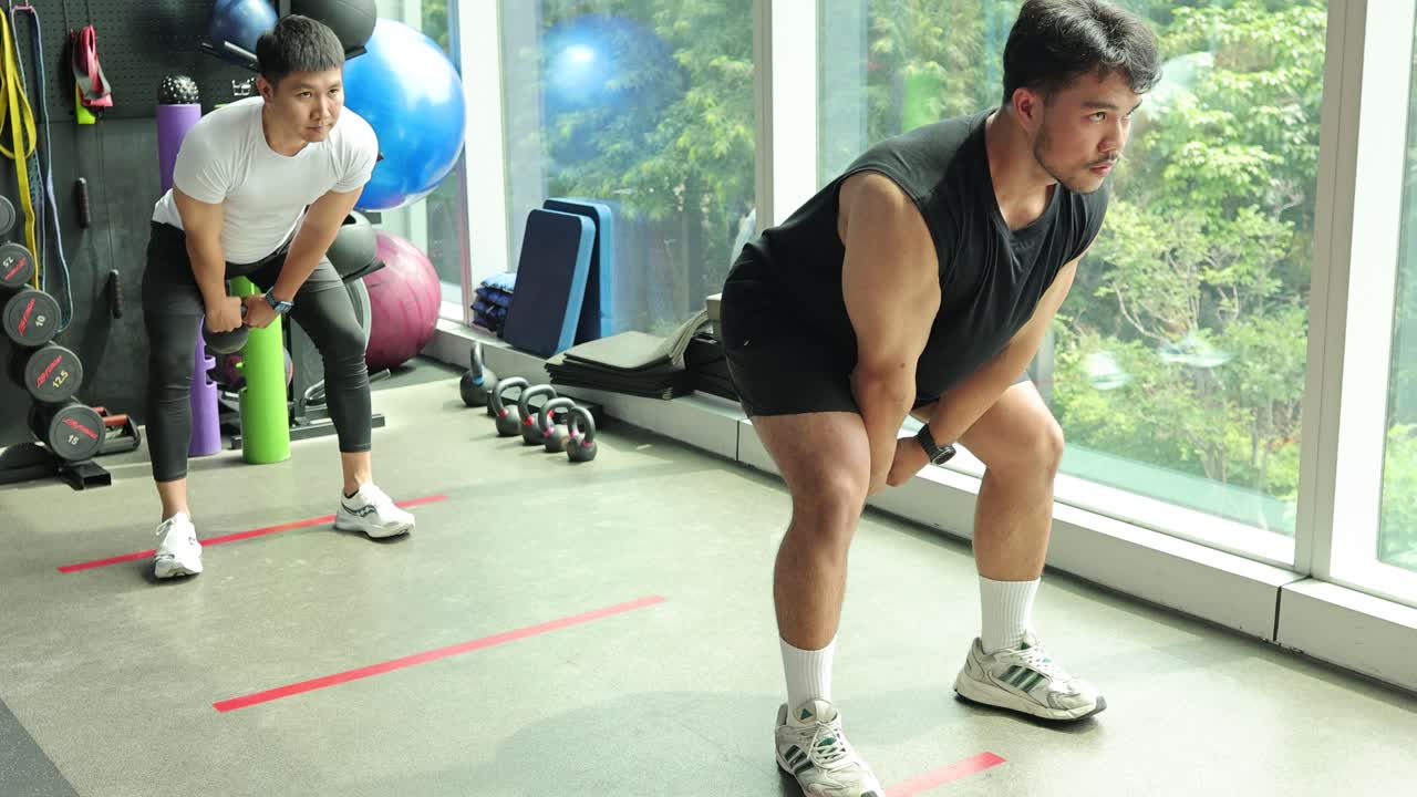 Two individuals perform kettlebell swings in a well-lit gym with large windows, focusing on strength and coordination