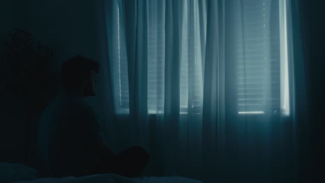 man sitting next to bed staring out at closed window blind shades, scratching head