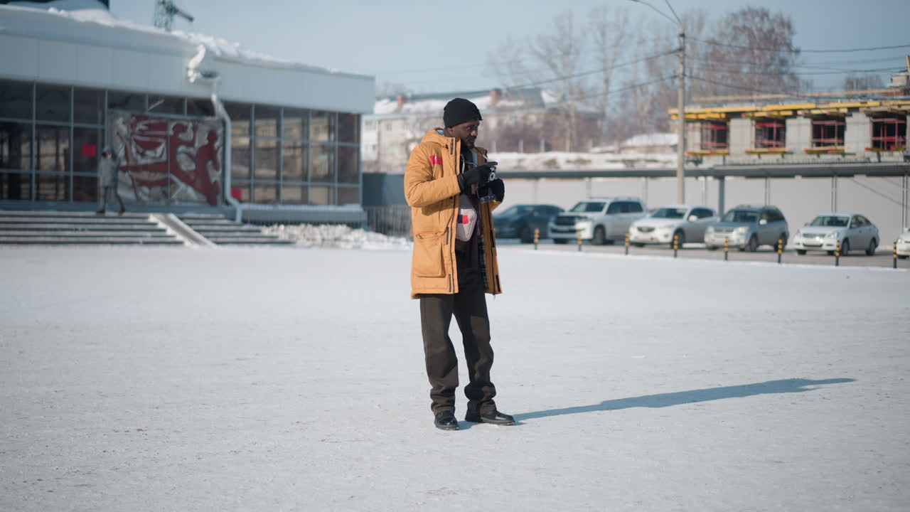 Campo lungo di un fotografo paesaggista con cappotto invernale, berretto, guanti e stivali robusti, in piedi su un'area urbana innevata sotto una luce diurna nitida, mentre scatta una foto dell'ambiente con la tracolla della macchina fotografica.