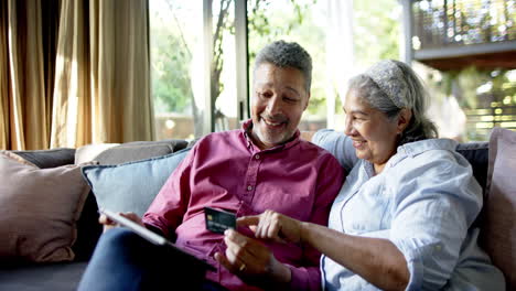 una pareja de ancianos feliz y diversa sentada en el sofá y usando una tableta para comprar en línea en casa