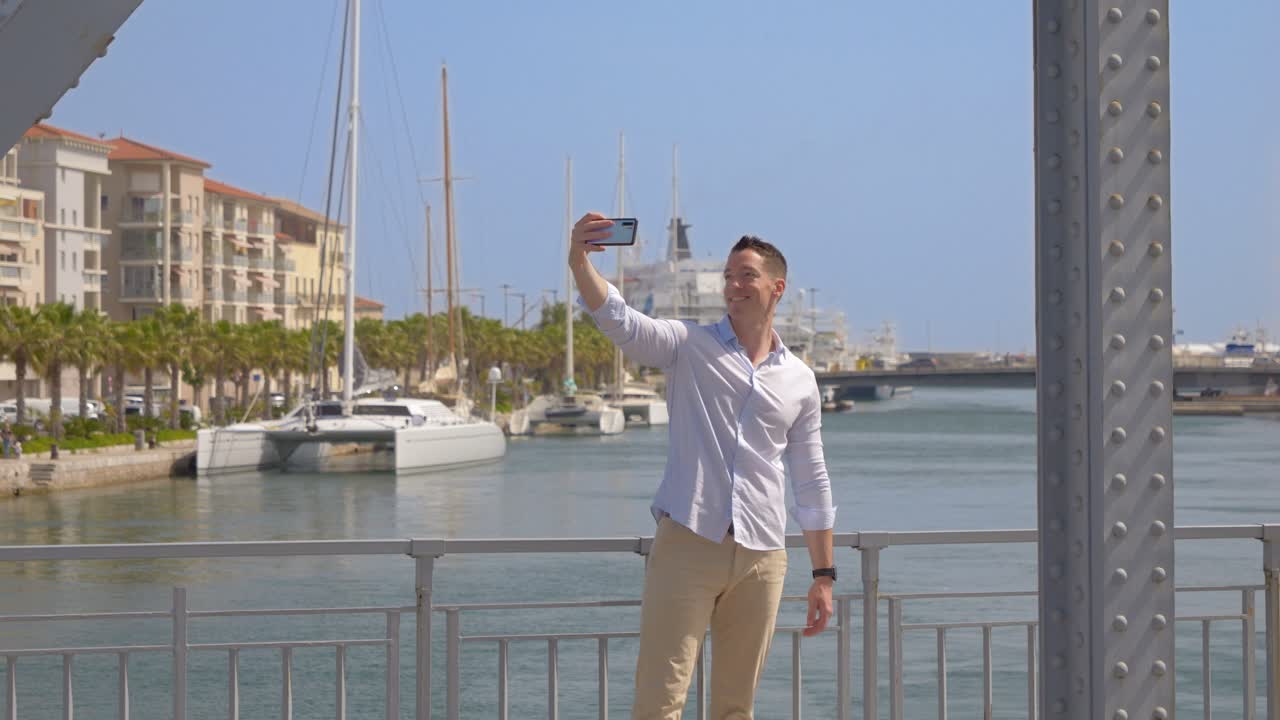 un giovane felice scatta dei selfie su un ponte di metallo con le macchine che passano, in una città soleggiata vicino al mare, con un catamarano sullo sfondo