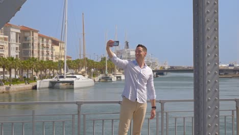 a happy young man takes selfies on a metal bridge with passing cars, in a sunny city by the sea, with a catamaran in the background