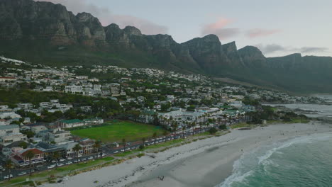 Vorwärts-Fliegen-über-Meeresküste,-Wellen-Waschen-Sandstrand.-Gebäude-In-Tropischem-Urlaubsziel,-Berge-Im-Hintergrund.-Kapstadt,-Süd-Afrika