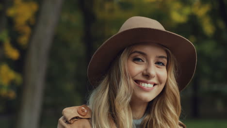 Close-up-view-of-caucasian-young-blonde-woman-in-a-hat-smiling-and-waving-at-camera-in-the-park-in-autumn