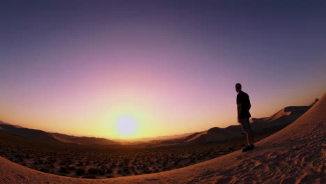 A man stands on a sand dune, admiring the sunset over a vast desert landscape
