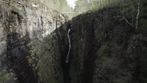 Drone-shot-flying-away-from-big-waterfall-inside-narrow-and-rocky-Corrieshalloch-gorge-in-Scottish-Highlands