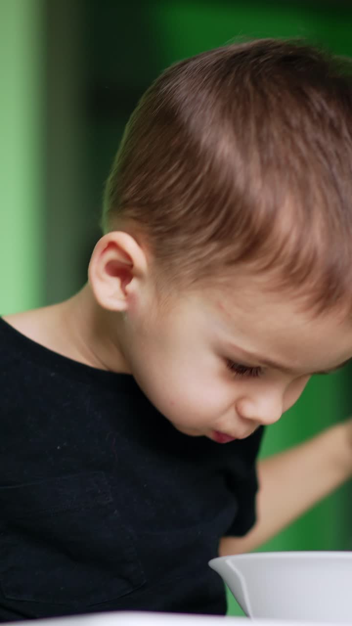 Adorabile bambino seduto davanti al piatto. Il bambino agita le mani allegramente durante i pasti. Inquadratura dal basso. Sfondo sfocato. Video verticale.