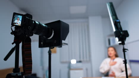 female blogger influencer speaks to camera. professional camera and daylight lamp are at foreground. blurred backdrop.