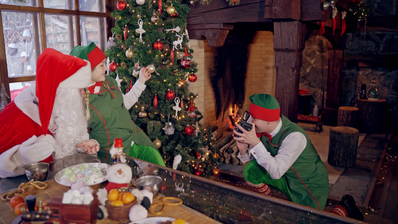 Santa with elf near the Christmas tree. Happy elf taking photo by retro camera on burning fireplace background. Happy xmas holidays.
