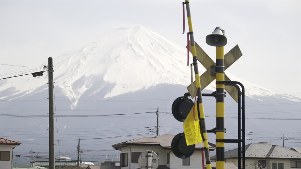 A railway crossing stands in the foreground as snow-draped Mount Fuji rises majestically in the background on a clear winter day. Nearby homes dot the landscape. Translation on flag ''do not cross''