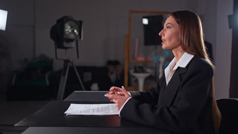 confident long-haired lady in black jacket sits at desk. reporter talks to camera in the light of soffits.