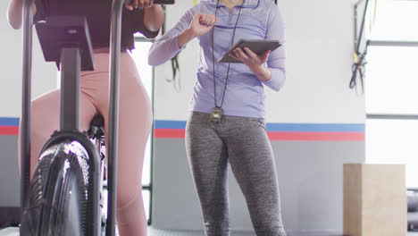 Video-De-Una-Entrenadora-Femenina-Diversa-Con-Tableta-Y-Una-Mujer-En-Bicicleta-Estática-Hablando-En-Un-Gimnasio