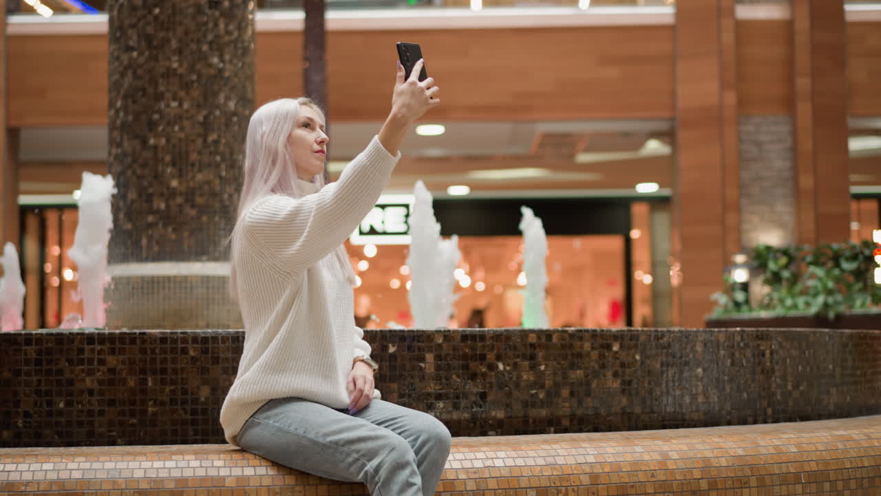 Elegant female content creator sits by indoor mall fountain raising mobile phone to photograph flowing water on serene mosaic bench as ambient shoppers stroll by, candid social media moment