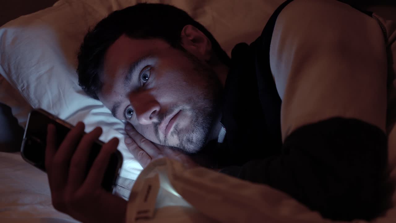hombre acostado en la cama con un teléfono inteligente, la pantalla ilumina la cara en un dormitorio oscuro, desplazándose, redes sociales, enviando mensajes de texto a amigos en la noche antes de dormir
