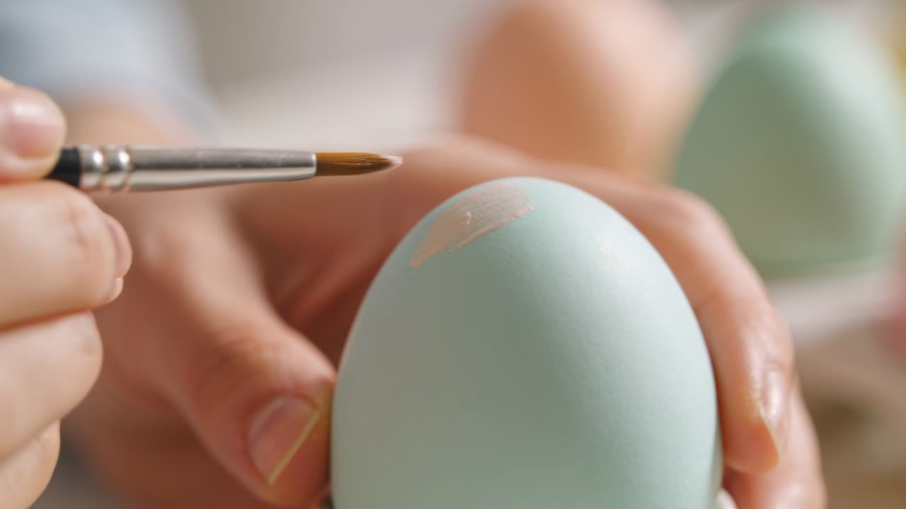 Painting hands in long sleeve turning pastel blue egg at home as brush contacting shell, decorating