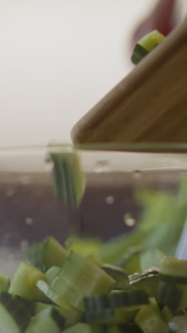 small cucumber pieces fall down into glass bowl from wooden board at cooking fresh salad on table in light kitchen extreme close view slow motion