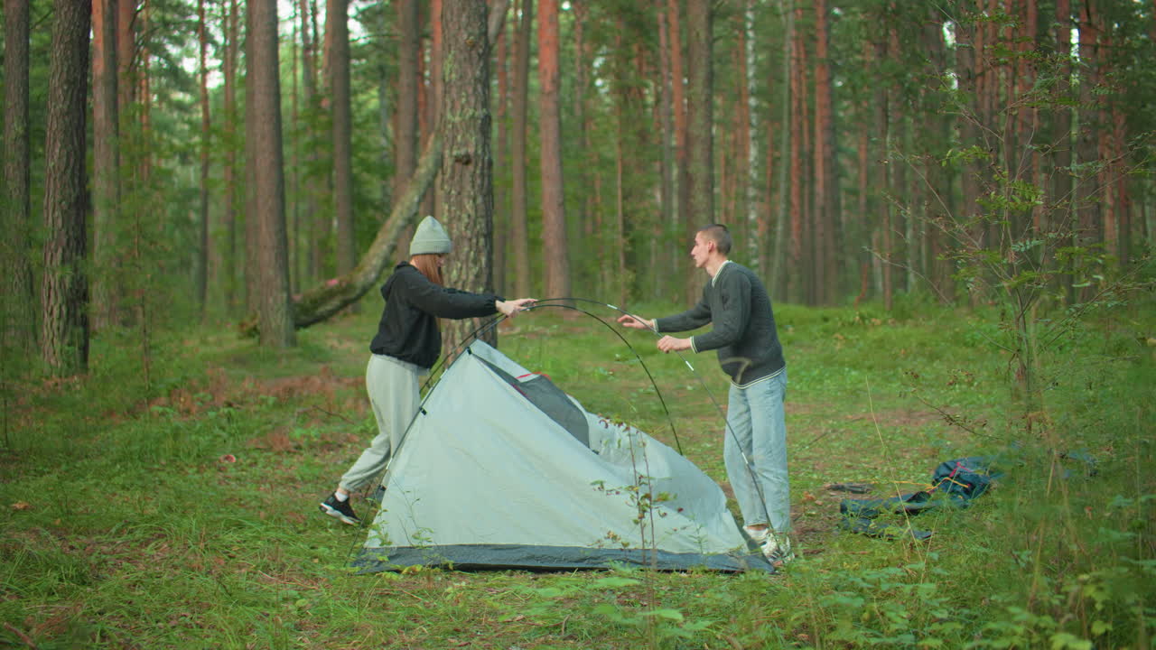 young man removes tent fabric while holding flexible pole as woman detaches her side during morning hour surrounded by tall forest trees and lush greenery in peaceful outdoor camping setting