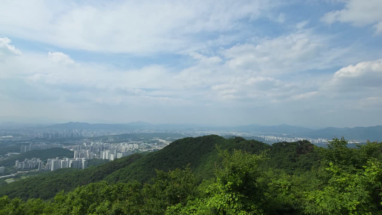 A stunning timelapse from Namhansanseong Mountain shows clouds drifting across a blue sky above the sprawling Seoul cityscape, with lush green forest covering the mountainside in the foreground.