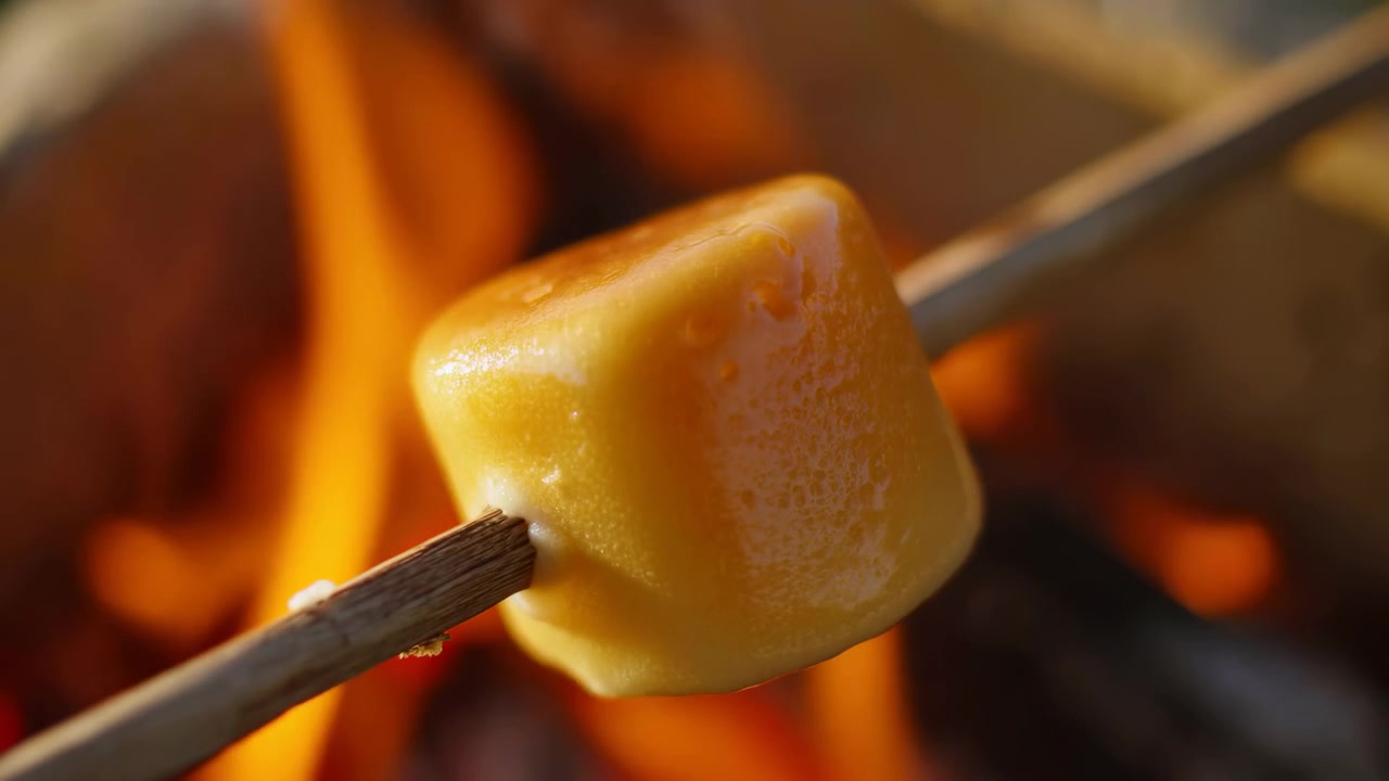 Vista ravvicinata di un marshmallow che gira su fiamme arancioni mentre i lati arrotondati si ammorbidiscono mostrando un colore tostato più intenso, con il fuoco ardente e lo sfondo sfocato che incorniciano il cibo