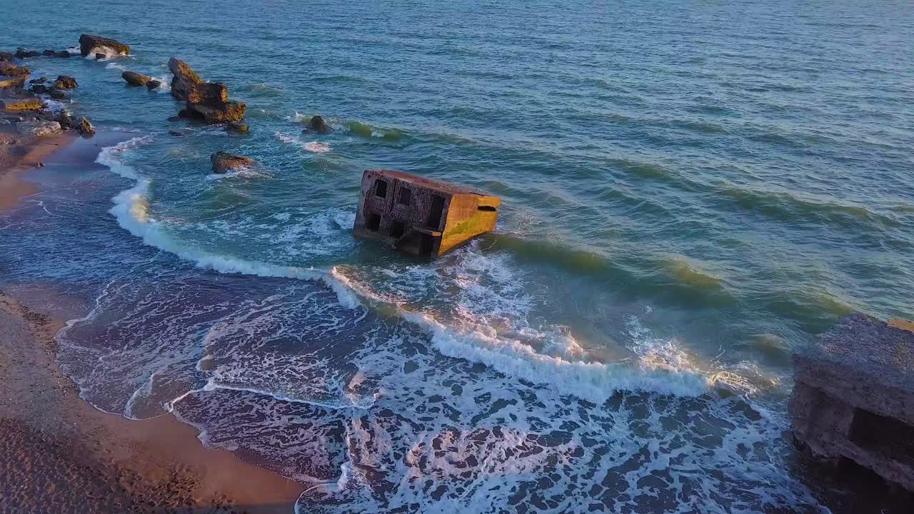 Luftfoto fra fugleperspektiv af forladte kystbefæstningsbygninger ved Karosta Nordlige Forts på Østersøens strand, bølger slår ind, solnedgang i den gyldne time, droneoptagelse der bevæger sig fremad
