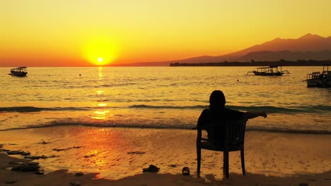 Silueta-De-Mujer-Joven-Sentada-En-Una-Silla-Sobre-Los-Flujos-Del-Mar,-Disfrutando-De-La-Romántica-Puesta-De-Sol-Con-Colores-Dorados-Que-Se-Reflejan-En-La-Tranquila-Superficie-Del-Mar-En-Bali