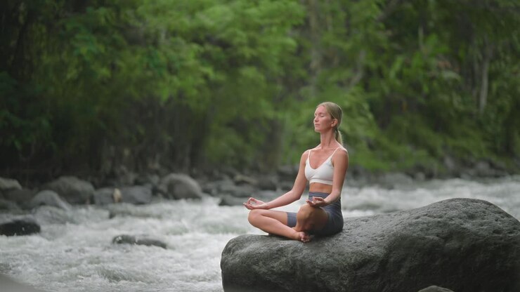 Mujer caucásica practicando la postura Sukhasana en una roca con un río caudaloso.