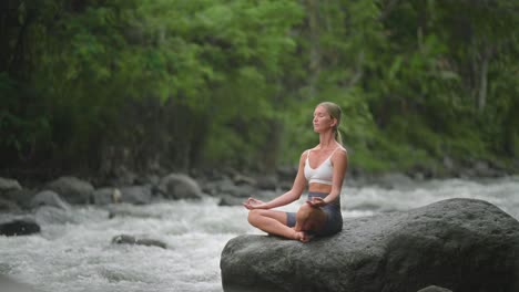 Hübsche-Kaukasische-Frau,-Die-Sukhasana-pose-Auf-Felsen-Mit-Fließendem-Fluss-übt