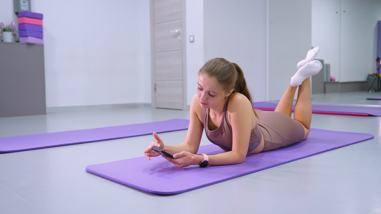 joven yace en la alfombra de yoga centrada en el teléfono en un moderno estudio de fitness, el espejo refleja su postura relajada y el entorno, creando una atmósfera serena