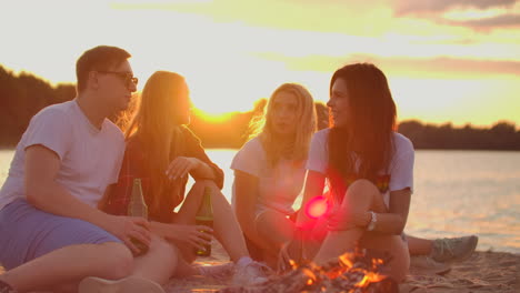 jóvenes estudiantes están sentados alrededor de una fogata en la playa con cerveza. están hablando entre sí al atardecer y disfrutando de la noche de verano en la costa del río.