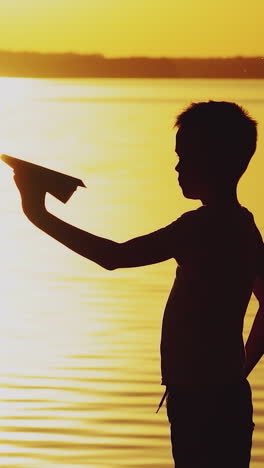 a little boy is holding a paper airplane in his hand and playing with him on the bank of the river at sunset on the background of the solar track. vertical video