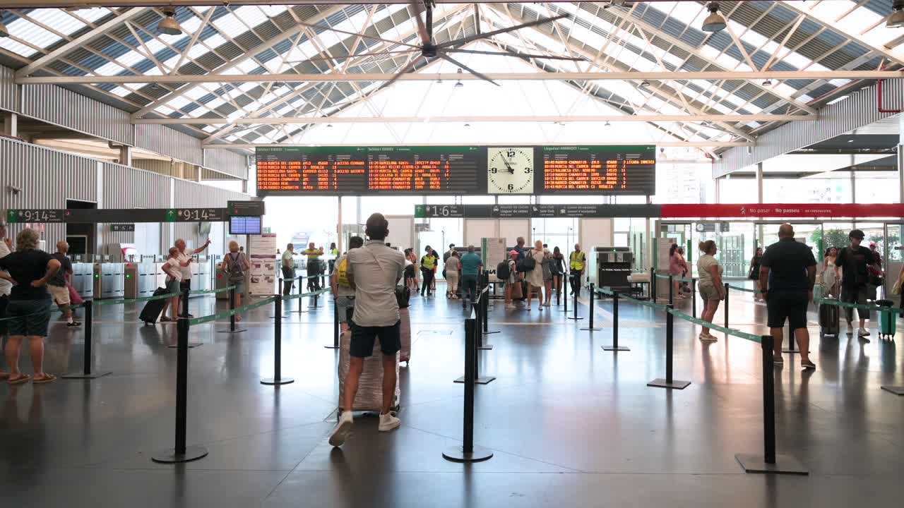 Wide shot of passengers arriving at the main terminal hall of Alicante train station and going through luggage security checks in Alicante, Spain.