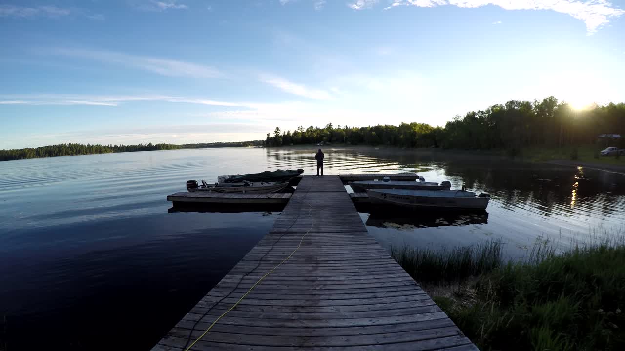 Timelapse from shore at Caliper Lake in Northwest Ontario, Canada. A man fishes on a dock centered in frame as the sun rises over calm water with clouds moving across the sky
