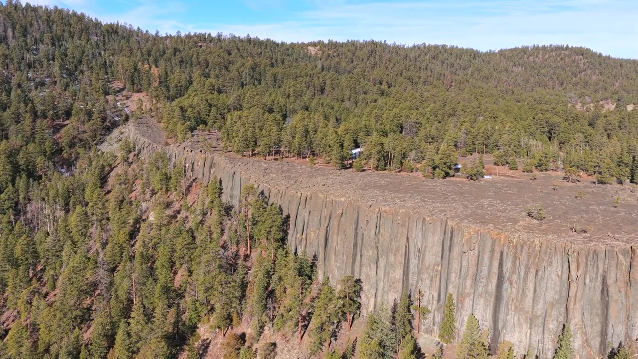 Prospettiva drone 4K di una mesa dalla cima piatta delimitata da un'imponente scogliera di basalto colonnare. Le fratture verticali della roccia vulcanica grigia si affacciano su una fitta foresta sempreverde nella natura selvaggia del Sud-Ovest