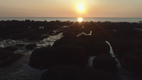 Very-Low-Aerial-Drone-Shot-Over-Silhouetted-Rocks-on-Beach-Flying-Forward-to-Calm-Sea-with-Small-Waves-at-Beautiful-Golden-Hour-Sunset-in-Old-Hunstanton-North-Norfolk-UK-East-Coast