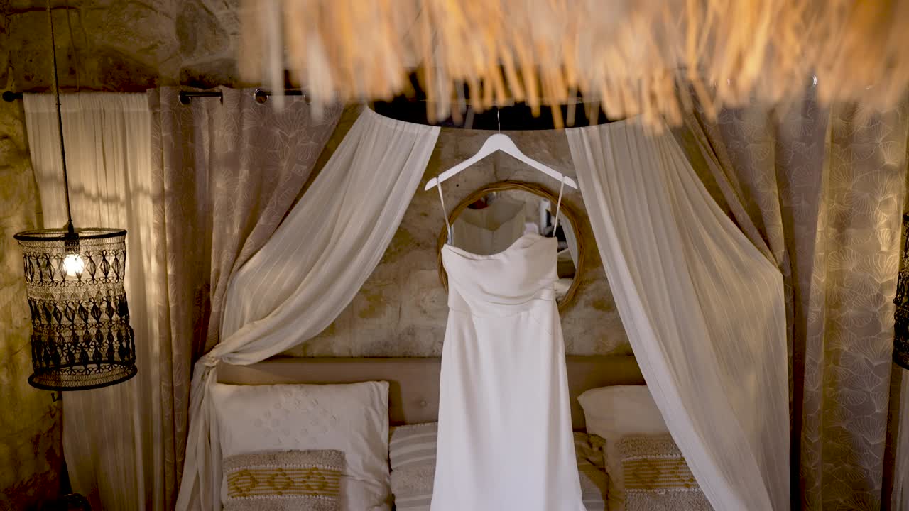 A stunning white wedding dress hangs gracefully in an inviting room, surrounded by warm lighting and elegant furnishings, perfect for a memorable occasion