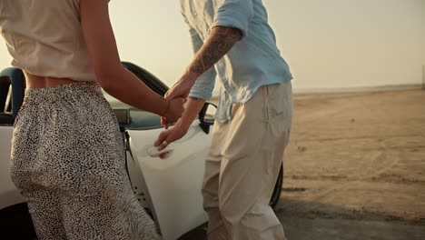 un tipo con un tatuaje en el brazo en una camisa azul abre la puerta de un coche descapotable blanco para su novia rubia durante el tiempo ventoso contra el río y un cielo amarillo