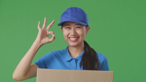 close up of asian female courier in blue uniform showing okay gesture and smiling while delivering a carton on green screen background in the studio