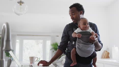 Father-With-Baby-Son-In-Sling-Multi-tasking-Working-From-Home-On-Laptop