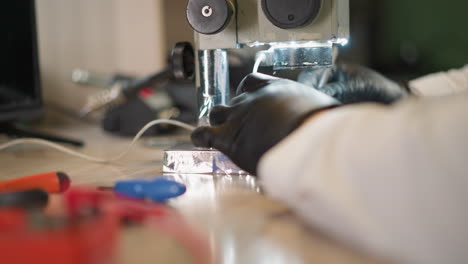 una vista de primer plano de un técnico con una bata de laboratorio blanca y guantes de mano negros trabajando en un circuito bajo un microscopio, con varias herramientas en la mesa con un fondo borroso