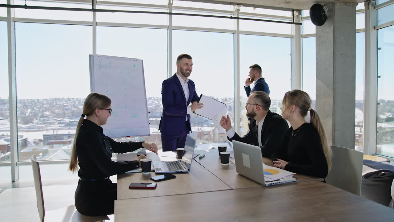 Colleagues sitting at the table in front of laptops and listening to the speaker. Male partner standing at the window and speaking on the phone at the backdrop.