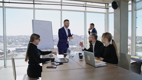 colleagues sitting at the table in front of laptops and listening to the speaker. male partner standing at the window and speaking on the phone at the backdrop.