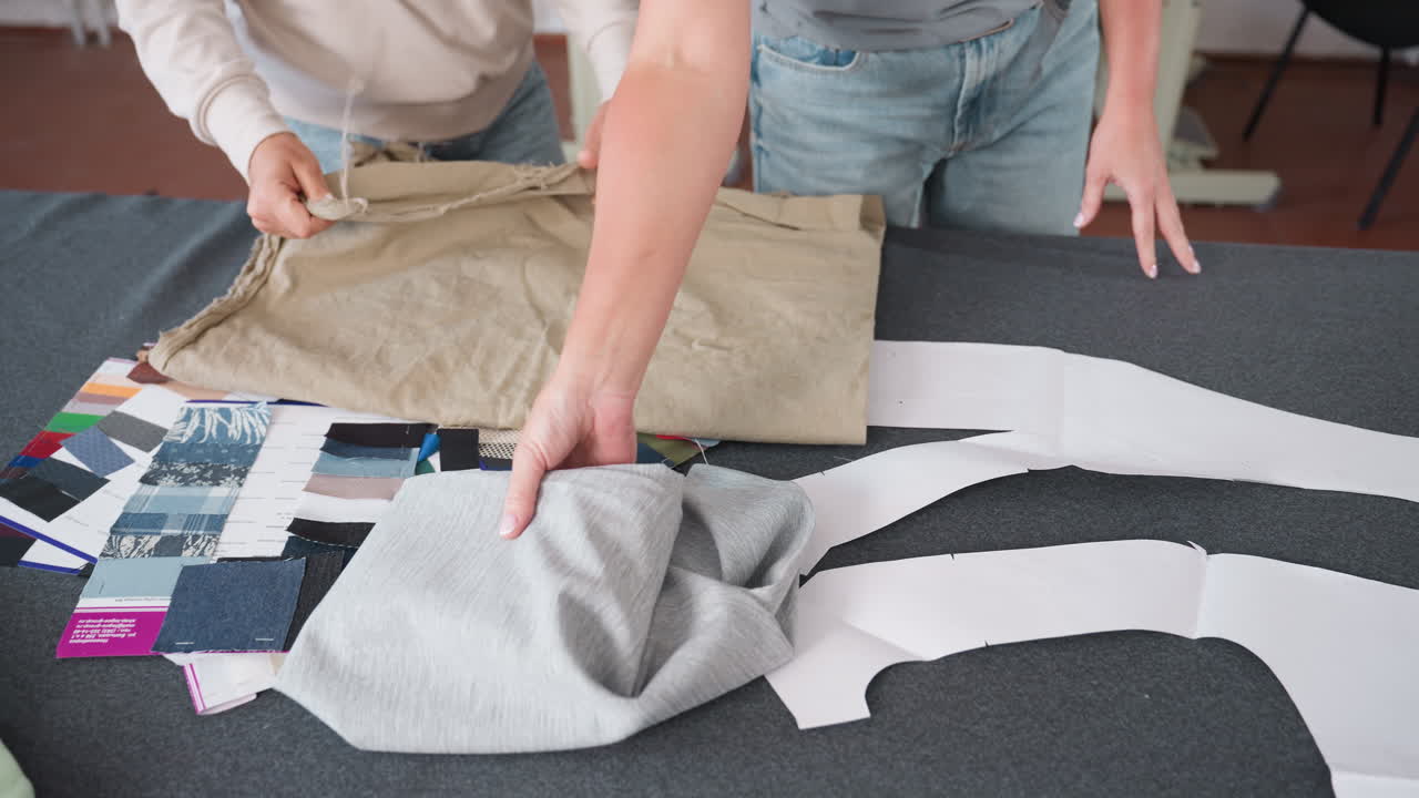 Female designers unfolding brown and ash fabrics on work table covered with colorful fabric samples and sewing patterns, preparing textiles for fashion project in creative workspace