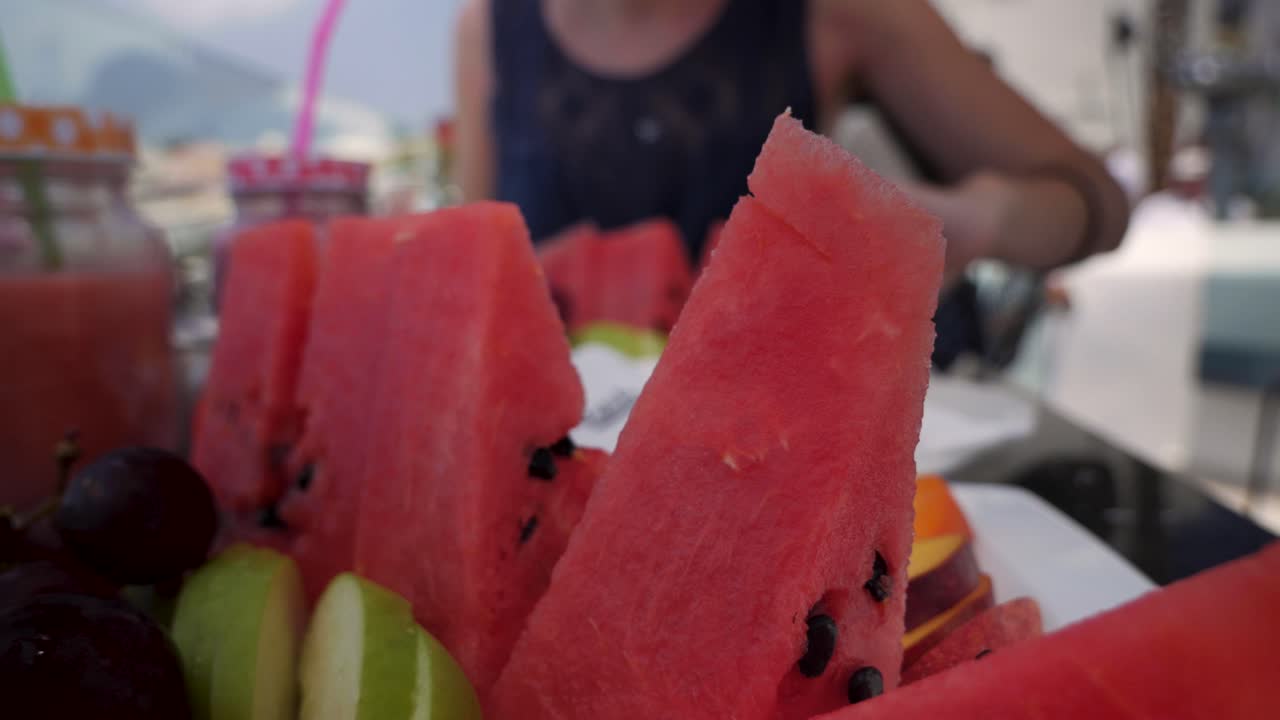 백그라운드에서 그녀의 전화를 가진 여자와 레스토랑에서 티라의 도시에서 산토리니 그리스에서 물 멜론, 사과, 포도와 과일 접시의 핸드 헬드 샷을 닫습니다