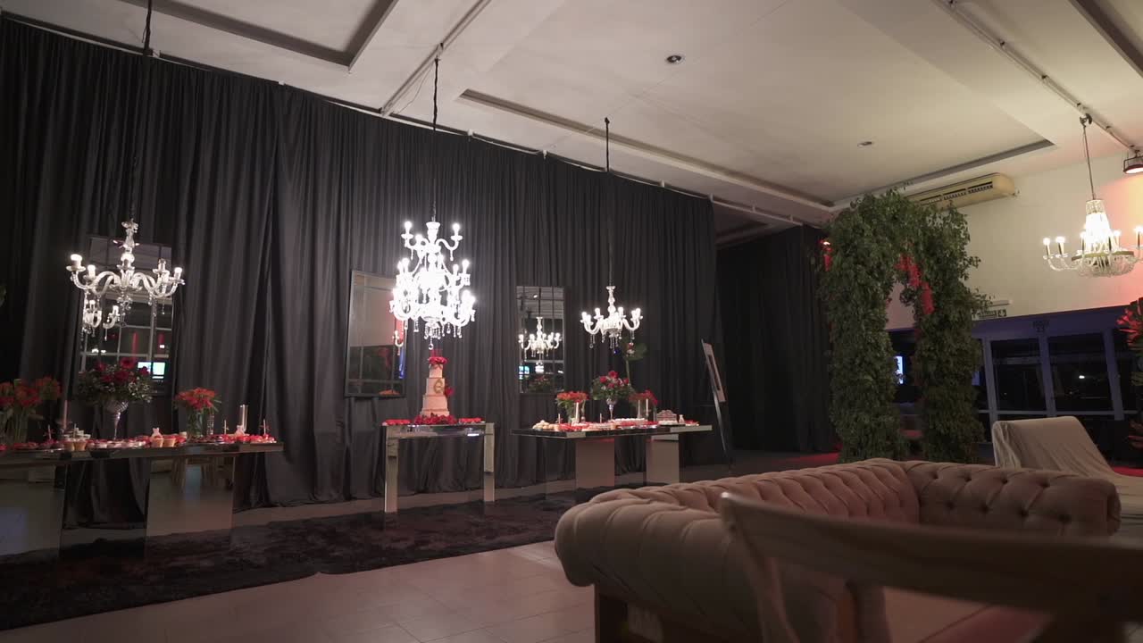 Lateral tracking general shot of spacious lounge room at birthday event, with classic brown couches, hanging white chandeliers, high white ceiling and floral arch arrangement. Shot on 1080p at 100fps.