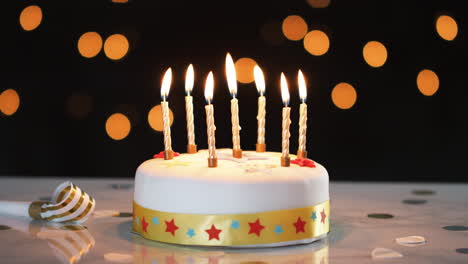 seven lit candles on a white, decorated birthday cake, a party blower beside it, bokeh lights in the background